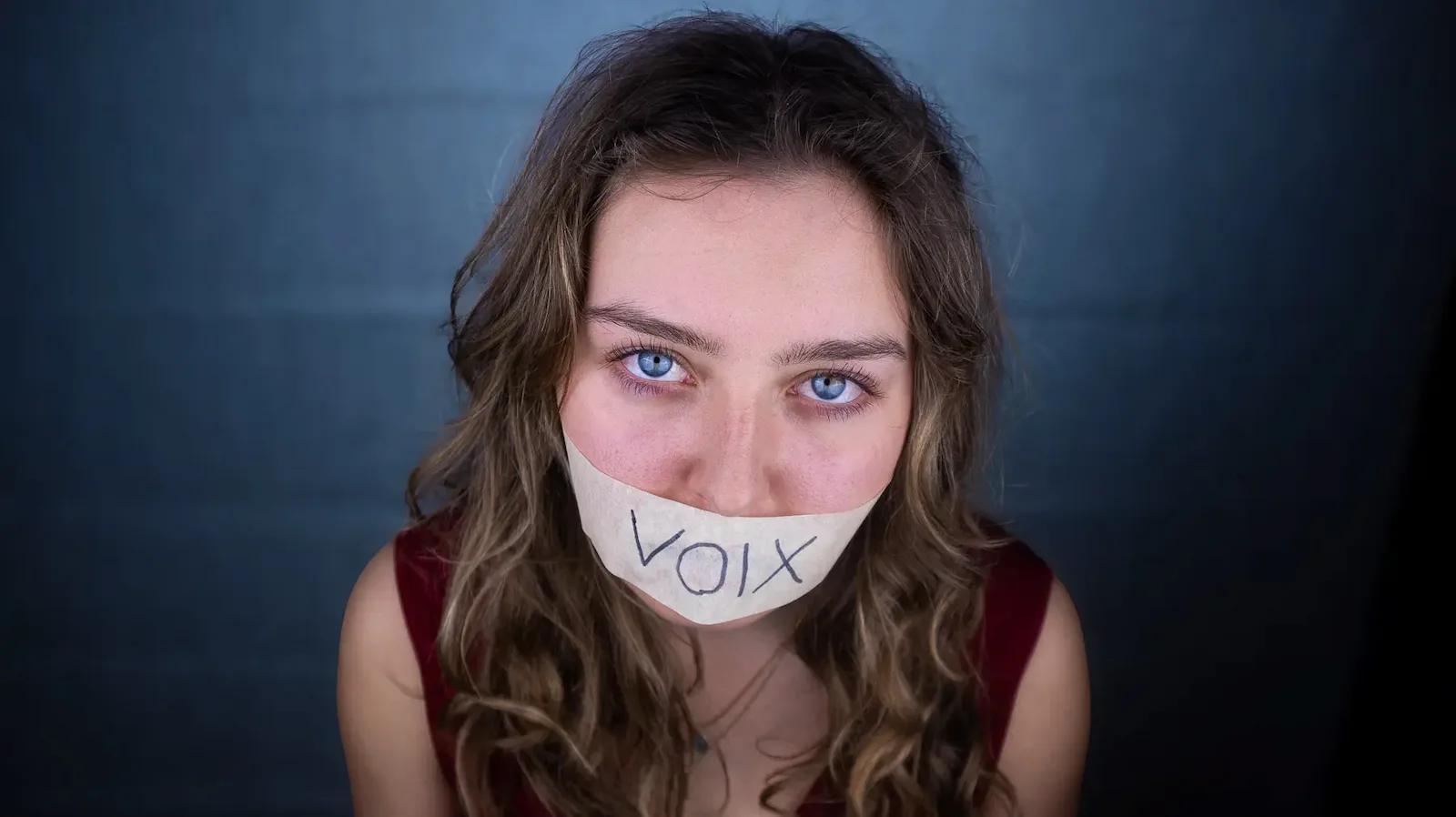 Portrait einer Frau mit Klebeband über dem Mund, auf dem „VOIX“ steht, die direkt in die Kamera blickt